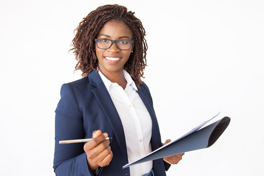 Happy successful business leader signing agreement, holding pen and documents, looking at camera. Young African American business woman posing isolated over white background. Signing contract concept