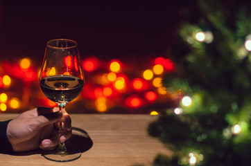 Hand toasting a glass of Rose wine on wooden table with Christmas tree and colorful bokeh light background.