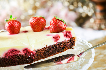 Delicious strawberries tart with vanilla cream on top. Chocolate cake and silver dessert scoop on glass tray with glitters in background.