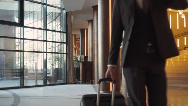 Young businessman in suit carrying a suitcase walking down the hotel lobby and talking on mobile phone, business trip.