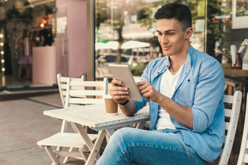 Outdoors Leisure. Stylish guy sitting at cafe on the street with cup of coffee using digital tablet...