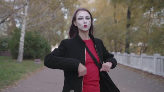 Mime girl in a black coat walks down the street showing joking performance. Steadicam shot.