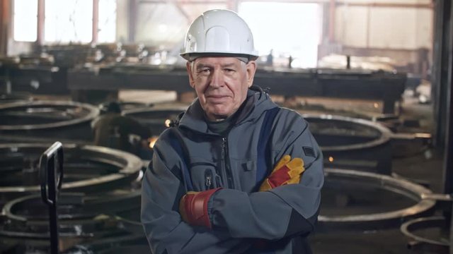 Panning portrait shot of cheerful elderly male worker in hard hat and gloves standing with folded arms in metal fabrication facility and smiling for camera