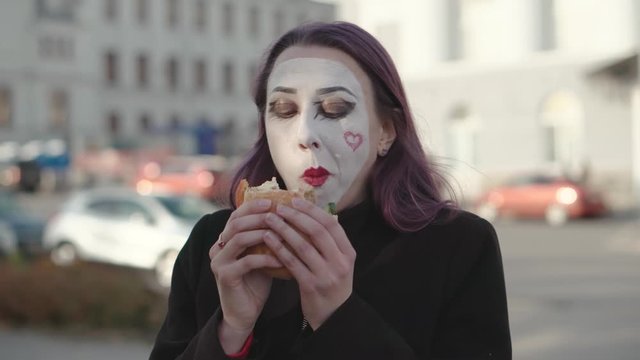 Mime girl in a black coat eats burger on the street.