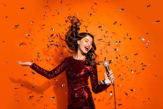Portrait of positive cheerful girl enjoy bachelor party celebration sing song in karaoke hold microphone wear burgundy red clothes isolated over orange color background confetti hair flying falling