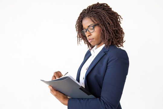 Focused accountant taking notes in paper, holding pen and folder, checking document. Young African American business woman posing isolated over white background. Paperwork concept