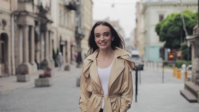 Portrait of Attractive Young Girl Wearing Fashionable Beige Trench Smiling and Laughing Looking to the Camera Standing at City Background.