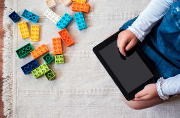 LIttle girl playing on her digital tablet on the floor near her colorful blocks.
