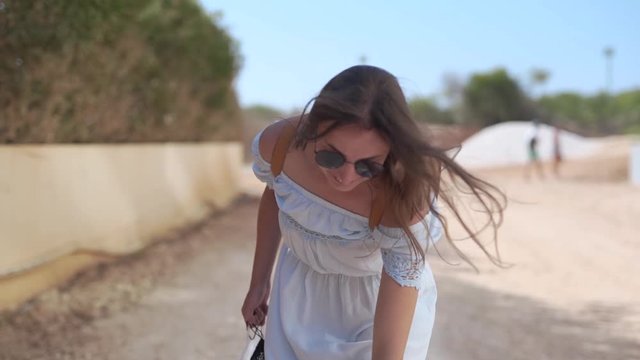 Beautiful girl is walking along the street on Cyprus. She is wearing a wide-brimmed hat and it is overthrowed with wind. Girl is smiling.
