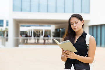 Fototapeta premium Concentrated young woman reading book. Young female college student with backpack standing on street and reading book. Education concept