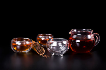 Glass tea cups, glass jug of tea, black tea in a scoop on a black background.
