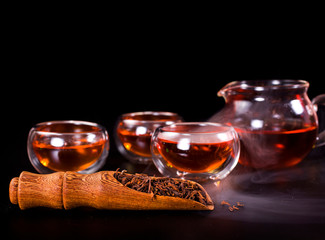 Glass tea cups, glass jug of tea, black tea in a scoop with vapour on a black background.