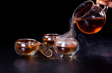 Chinese tea ceremony. Pouring of famous chinese puerh tea from a glass jug into a cup with the vapour. Still life on a black background.