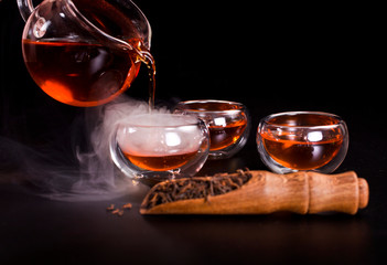 Chinese tea ceremony. Pouring of famous chinese puerh tea from a glass jug into a cup with the vapor. Still life on a black background.