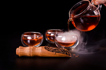 Chinese tea ceremony. Pouring of famous chinese puerh tea from a glass jug into a cup with the vapour. Still life on a black background.
