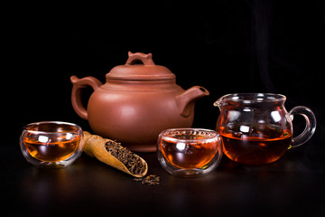 Clay tea pot, glass jug with cups of tea and dried black tea in a scoop on a black background.