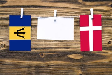 Hanging flags of Barbados and Denmark attached to rope with clothes pins with copy space on white note paper on wooden background.Diplomatic relations between countries.