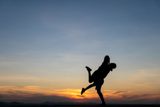 A silhouette of a happy young couple hugging, with the man lifting the woman off of her feet, at sunset.