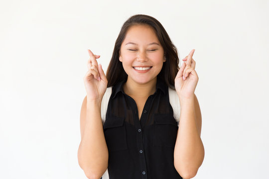 Excited student girl with closed eyes keeping fingers crossed. Young Latin woman in casual standing isolated over white background. Making wish concept