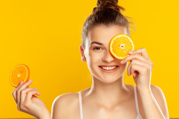 Young beautiful woman holding slice of orange in front of her eye on yellow background