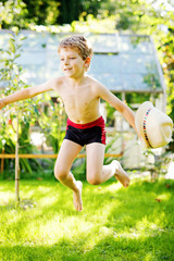 Cute active kid boy jumping in the garden on warm sunny summer day. Happy kid looking at the camera. Adorable child with blond hairs and blue eyes