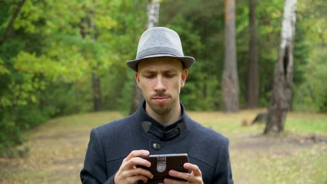 A man in a hat and coat in the rain looking at something on the phone
