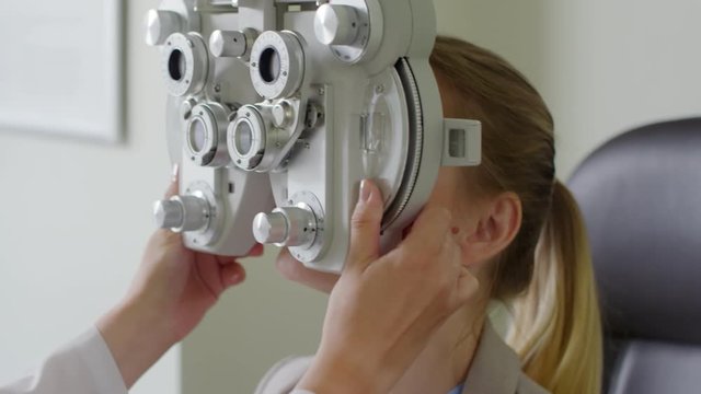 Tracking shot of smiling woman looking into phoropter instrument as cheerful female optometrist asking questions during eye exam