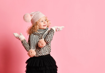Baby girl in warm hat, gloves standing with hands up holding catching something on open palms on pink with copy space