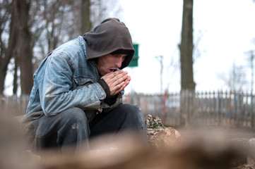Homeless, sad man in a beanie sitting outside and feeling cold, trying to warm up his hands