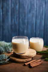 tasty eggnog cocktail on round wooden boards near spruce branches and cinnamon sticks on blue textured background