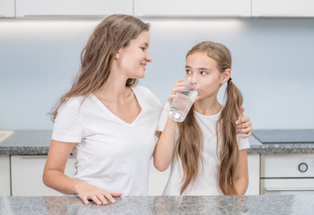 Young girl drinks water with happy mother at kitchen