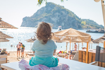 Fototapeta na wymiar Amazing view of little girl sitting on a table and looking on turquoise seaside in sunny summer day, Corfu, Greece
