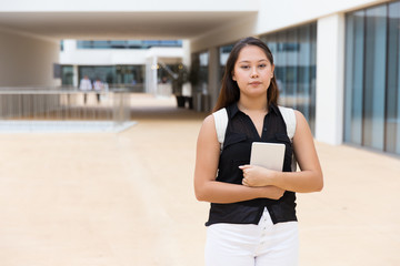 Serious confident female student embracing tablet. Young Asian woman walking outside, holding digital device, looking at camera. Student with gadget concept