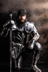 Fototapeta na wymiar handsome knight in armor holding sword and bend knee on black background
