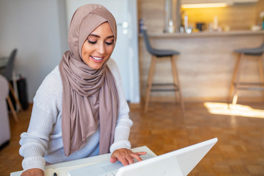 Beautiful Women Wearing The Islamic Headscarf, Sitting On A Wooden Table Holding Pencil Writing On Notebook And Cute Smile With Computer. Attractive Female Arabic Working On Laptop Computer