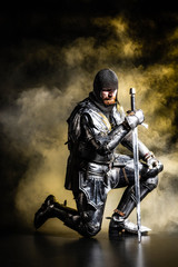 handsome knight in armor holding sword and bend knee on black background - obrazy, fototapety, plakaty