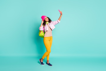 Full length body size photo of turned cheerful pretty cute nice charming girlfriend wearing yellow pants trousers pink cap headwear footwear jacket taking selfie isolated teal color background