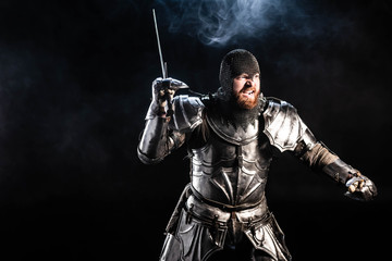 handsome and angry knight in armor fighting with sword on black background