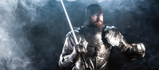 Fototapeta na wymiar panoramic shot of handsome knight in armor looking away and holding sword on black background