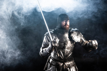 handsome knight in armor looking away and holding sword on black background