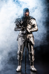 Fototapeta na wymiar handsome knight in armor looking at camera and holding sword on black background