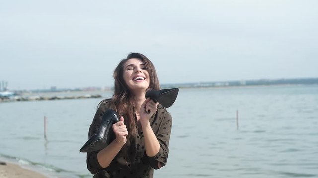 beautiful girl with dark hair in green dress is dancing merrily on sandy sea beach. woman holds slipper with heel in her hands and actively and rhythmically moves. sea tour. happy woman on vacation.
