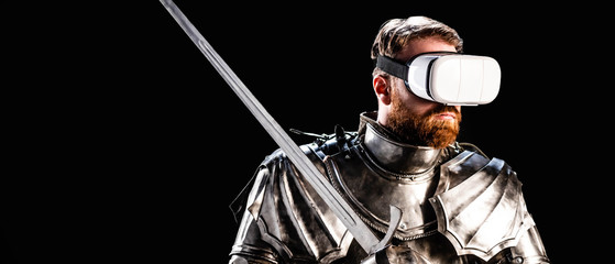 panoramic shot of knight with virtual reality headset in armor holding sword isolated on black - obrazy, fototapety, plakaty
