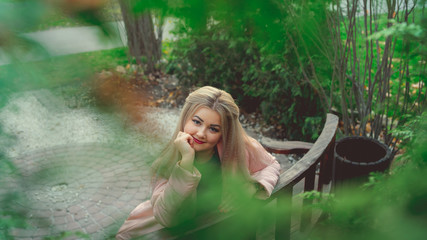 A portrait of a beautiful girl, sitting on a bench near the green vegetation.