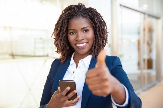 Happy Satisfied Female Customer Holding Cellphone, Making Like Gesture, Showing Thumb Up. Young African American Business Woman Standing Near Outdoor Glass Wall. Positive Feedback Concept