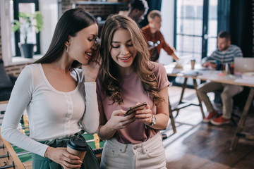 attractive, smiling businesswoman using smartphone near cheerful colleague holding coffee to go