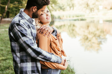 handsome man embracing happy girlfriend while standing near lake in park