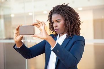 Focused female professional taking picture on cellphone outside. Young African American business woman standing near outdoor glass wall. Photo concept - obrazy, fototapety, plakaty