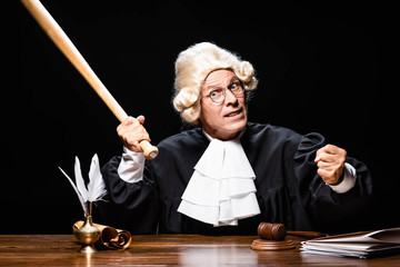 angry judge in judicial robe and wig sitting at table and holding bat isolated on black