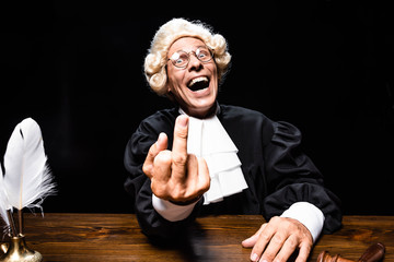 smiling judge in judicial robe and wig sitting at table and showing middle finger isolated on black